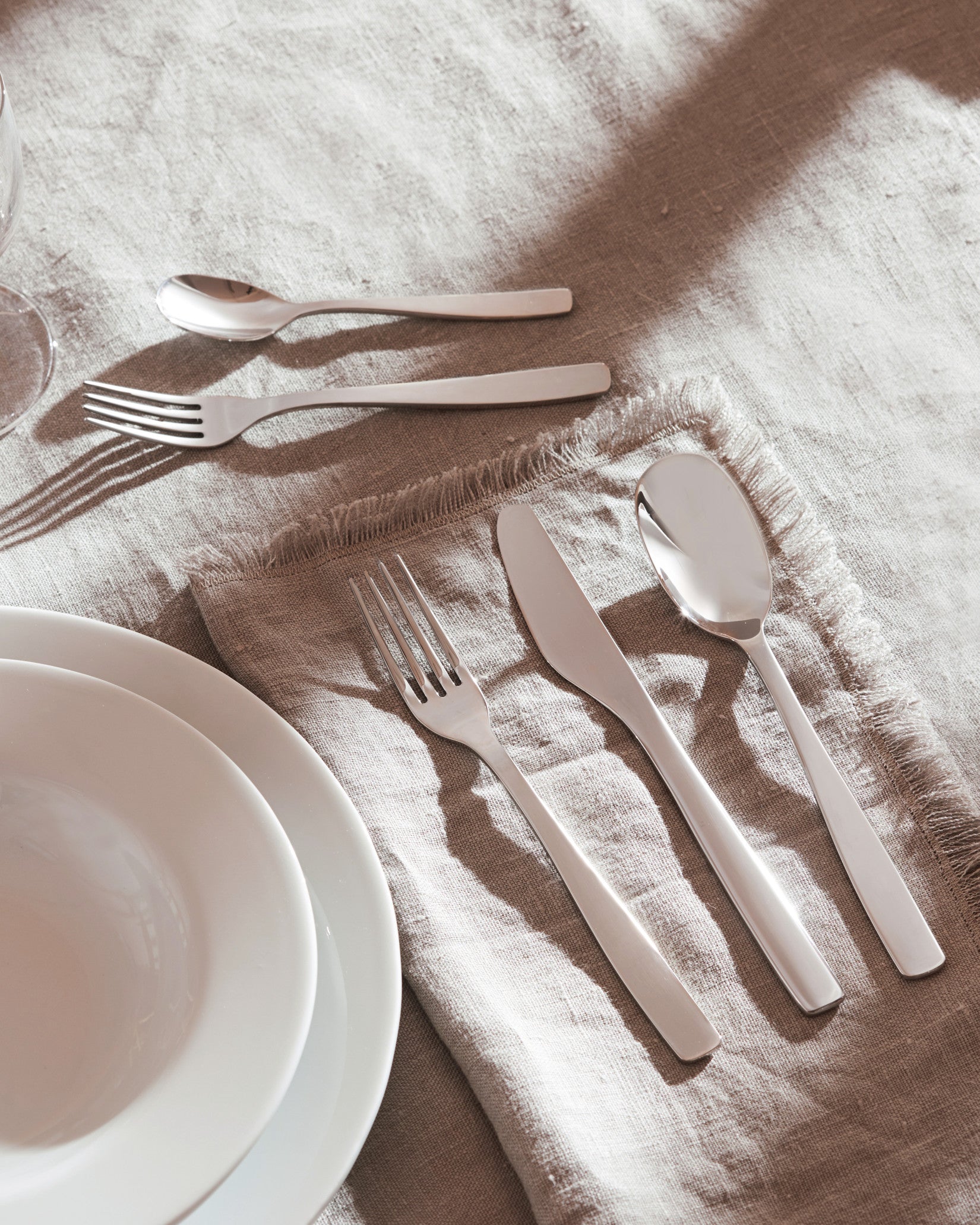 Silver polished cutlery set by Alessi, laid on a beige linen tablecloth with white plates and a glass, in bright sunlight.