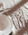 Silver stainless steel cutlery set by Alessi, laid on a fringed linen placemat with white plates, bathed in sunlight.