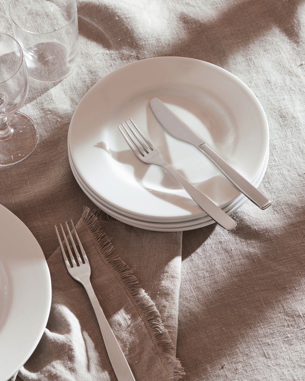 Silver-toned KnifeForkSpoon cutlery by Alessi, featuring a knife and forks on white plates and a fringed linen napkin with sun shadows.