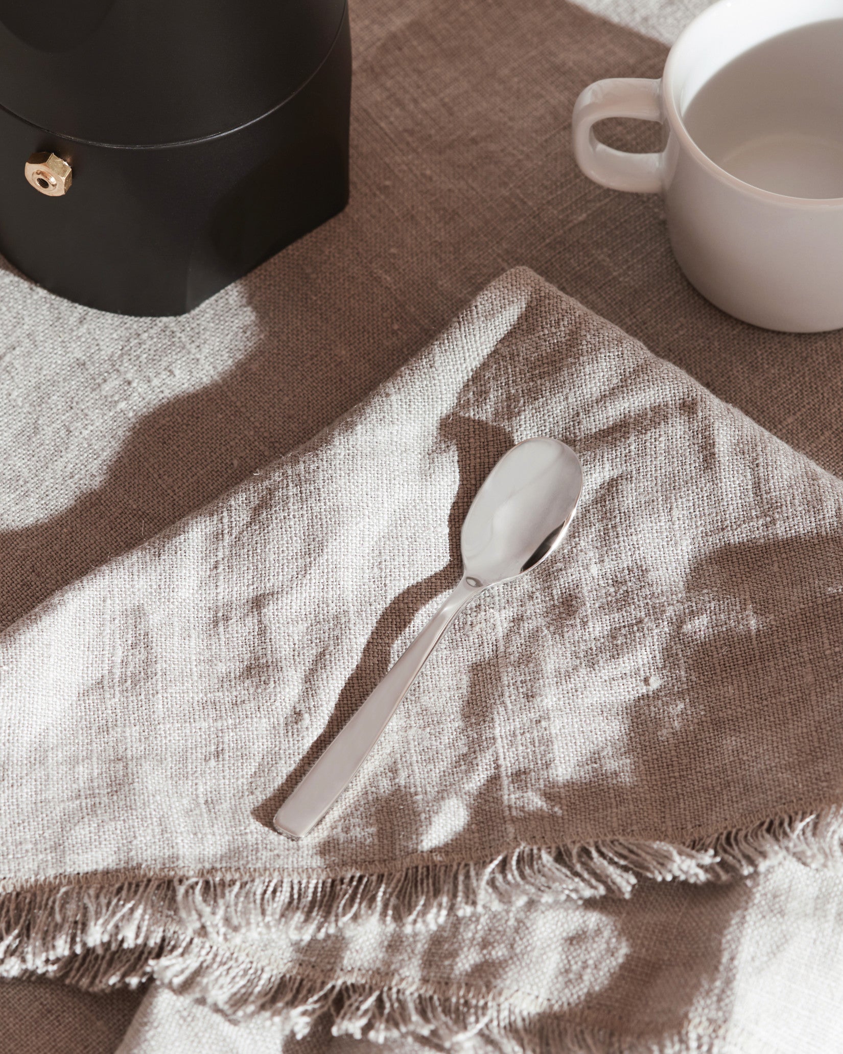 Silver spoon by Alessi, reflecting light on a textured linen napkin with fringe, alongside a black coffee maker and white mug.