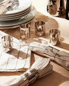 Silver-toned metallic napkin rings by Alessi with various playful cut-out figures, displayed on a wooden table with linen napkins.