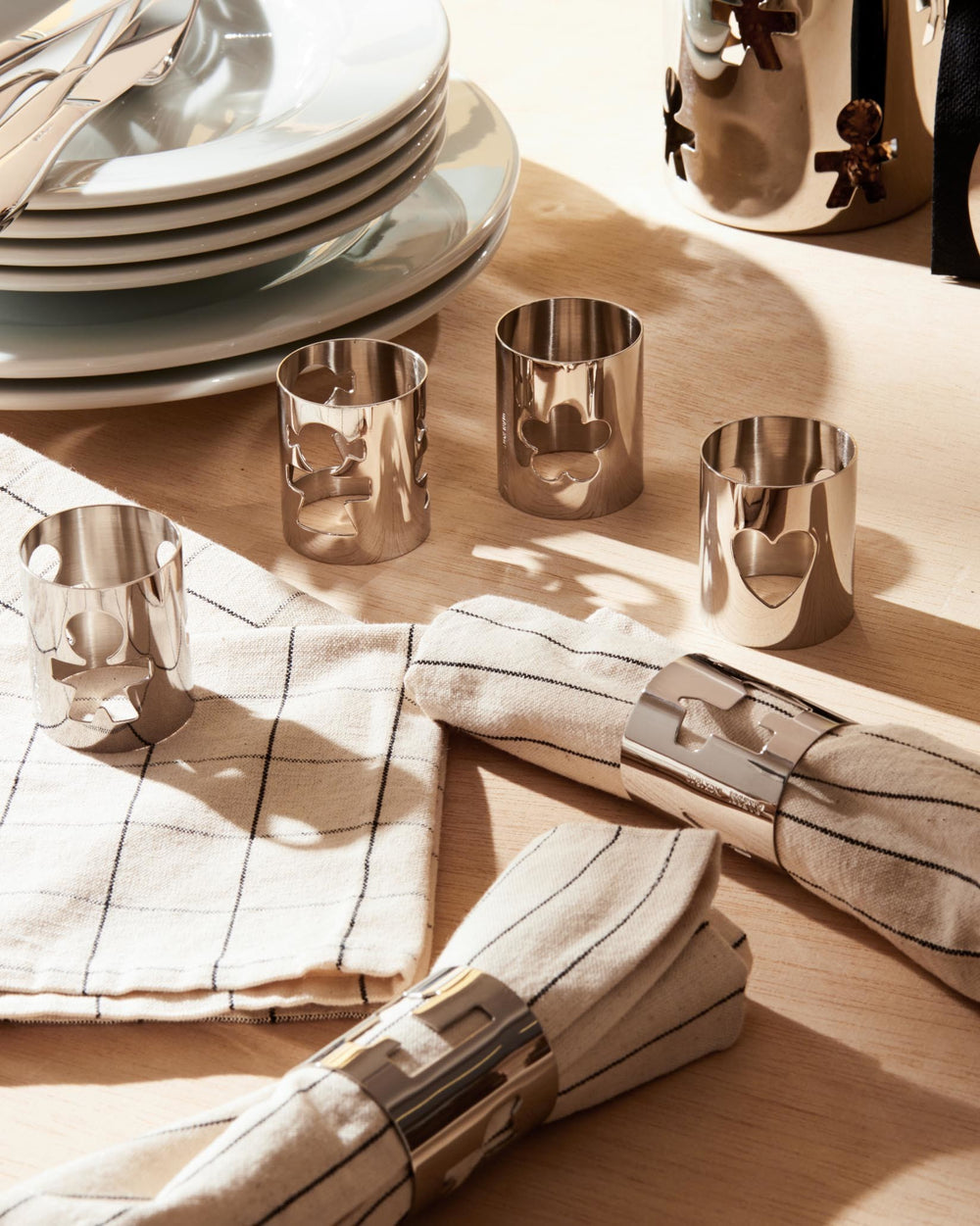 Silver-toned metallic napkin rings by Alessi with various playful cut-out figures, displayed on a wooden table with linen napkins.