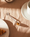 Silver-colored metal coffee spoon by Alessi with a smiling face handle, holding brown sugar on a beige linen tablecloth.