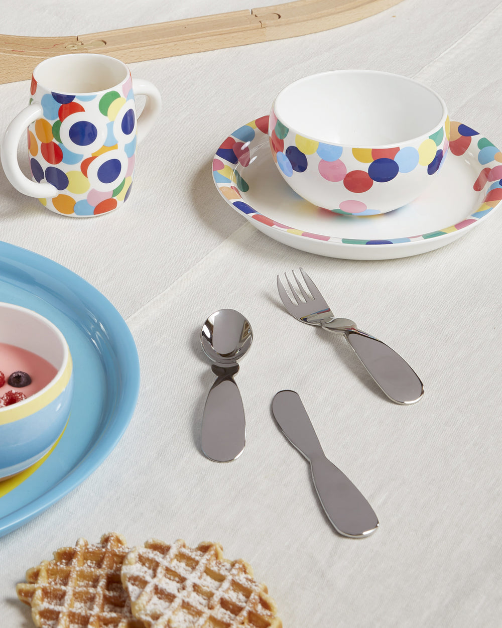 White multi-colored polka dot children's dinnerware by Alessi, with a two-handled mug, bowl, and plate, alongside child-sized cutlery on a white tablecloth.