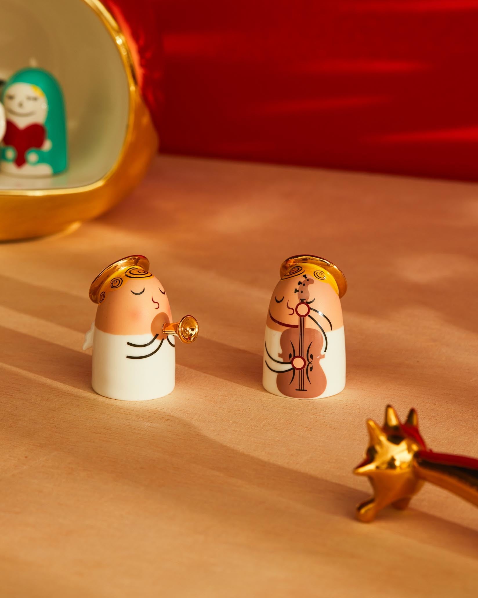Two white and peach angel figurines with golden accents by Alessi, one playing a trumpet and the other a cello, on a wooden surface.