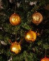Golden male character Christmas ornament by Alessi, with a stylized face and textured hair, hanging on a green Christmas tree.