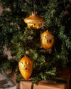 Golden character Christmas ornament by Alessi with crown and faceted gems, hanging on a green Christmas tree with gift boxes below.