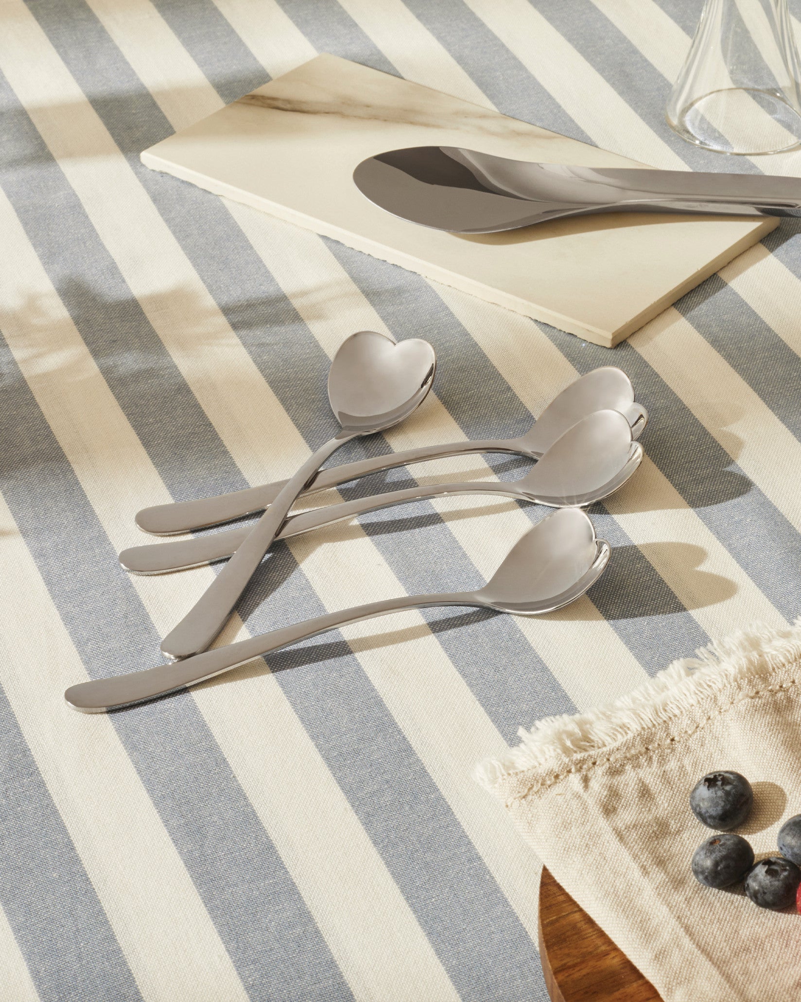 Silver heart-shaped spoons by Alessi, arranged on a blue and white striped tablecloth with a large serving spoon and blueberries.