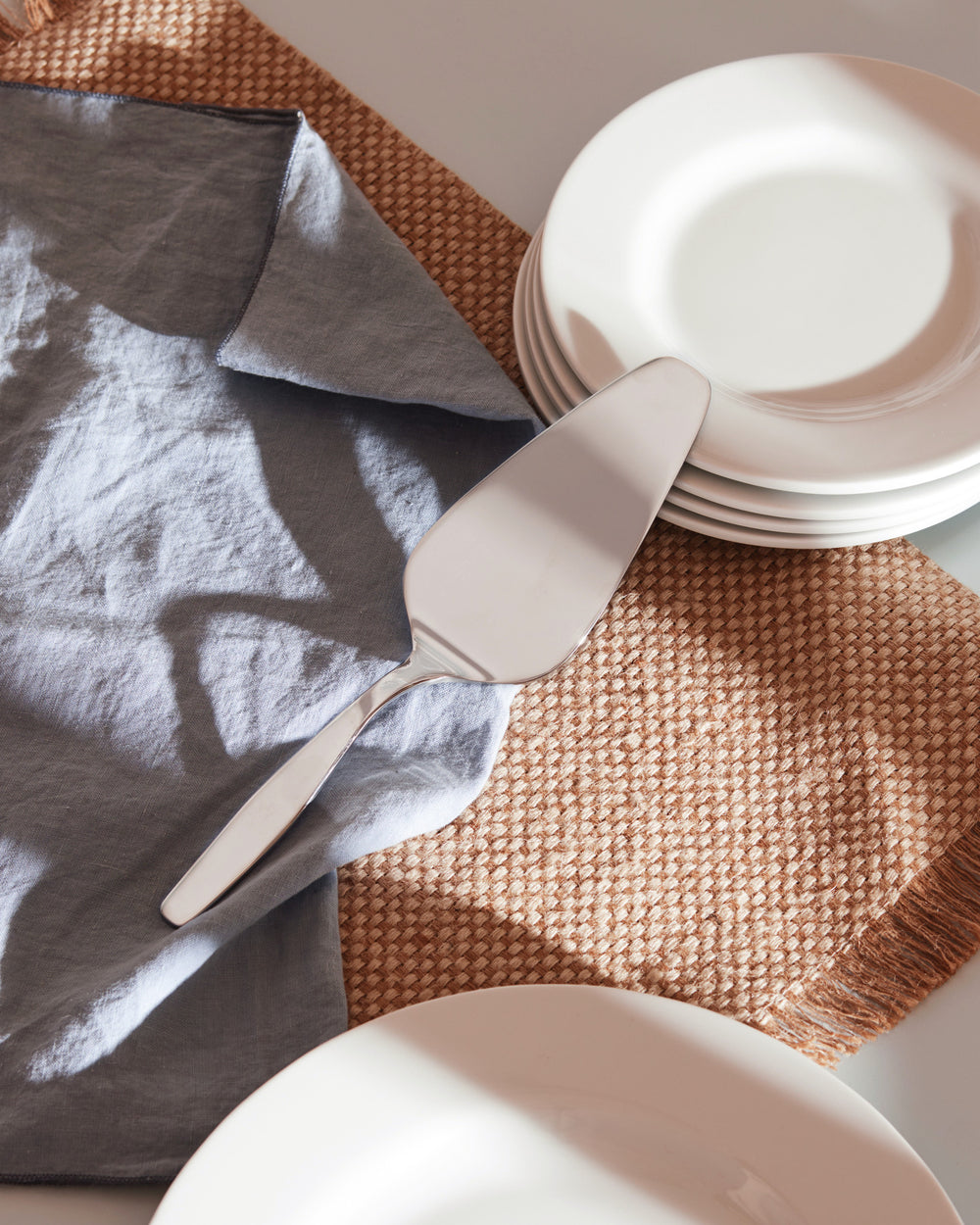 Angled silver metal cake server Itsumo by Alessi, on a blue linen napkin with white plates and a woven placemat.
