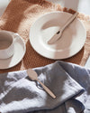 Silver metal spoon by Alessi, resting on a light blue linen napkin next to a white plate and cup.