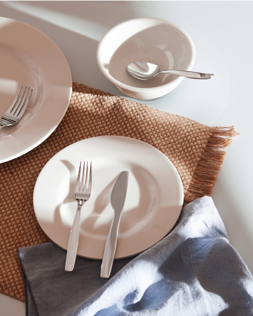 Polished silver cutlery by Alessi, featuring a fork, knife, and spoon on white plates with a woven placemat.