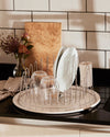 Round stainless steel dish rack with cream tray by Alessi, holding white plates and clear glasses on a black countertop.