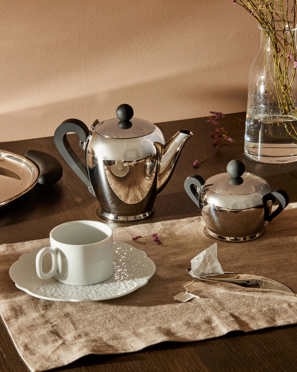 Polished chrome tea set with black handles by Alessi, featuring a teapot, sugar bowl, and white teacup.