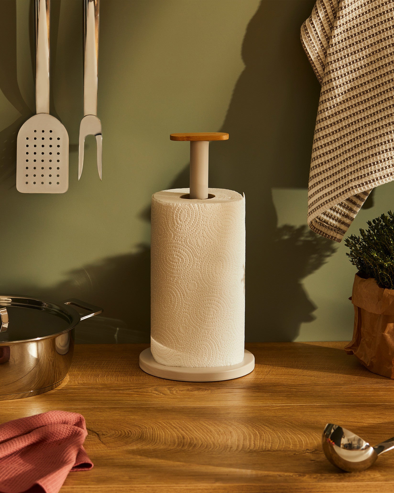 Matte beige and wood paper towel holder by Alessi holding a roll of white paper towels on a wooden countertop.