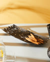 Dark brown elongated metal bread basket by Alessi, with intricate laser-cut bark patterns, filled with sesame breadsticks.