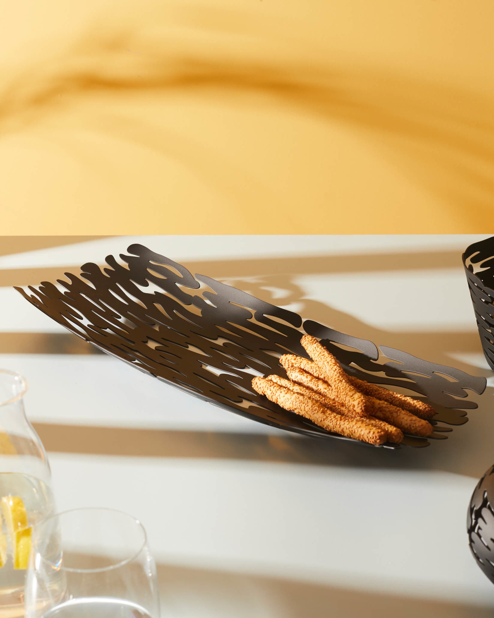 Dark brown elongated metal bread basket by Alessi, with intricate laser-cut bark patterns, filled with sesame breadsticks.