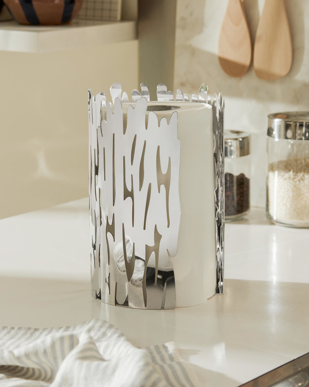 Silver metallic paper towel holder by Alessi featuring an organic cut-out design, holding a white roll in a kitchen.