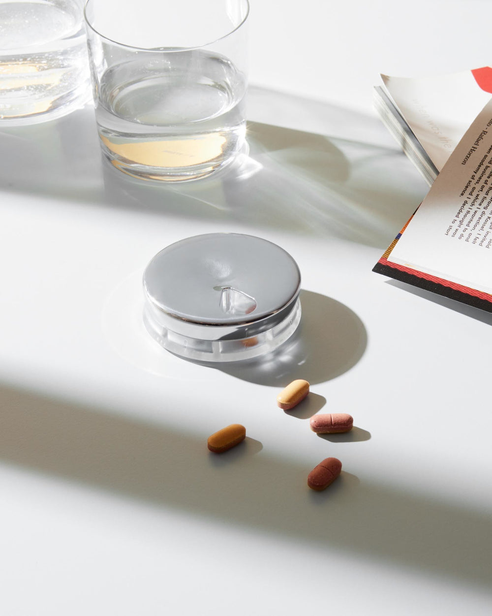 Silver and clear round pill organizer by Alessi, on a white table with water glasses and scattered pills.