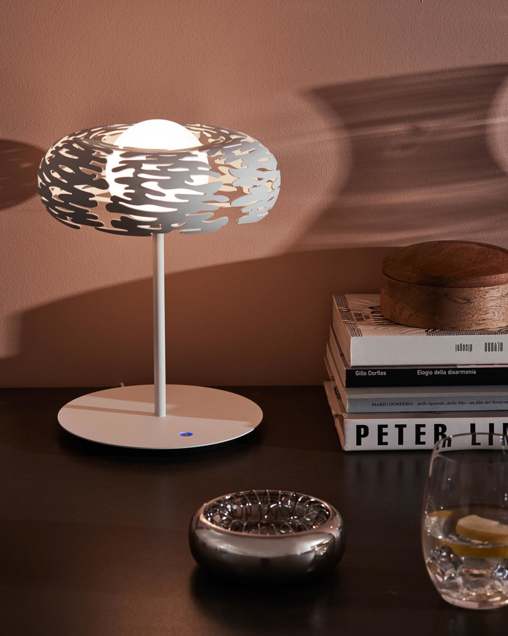 White table lamp by Alessi with an intricate, organic-patterned shade, casting shadows on a wall, styled on a dark surface.