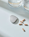 Polished silver chestnut-shaped object by Alessi, displayed with brown capsules and two water glasses on a blue surface.