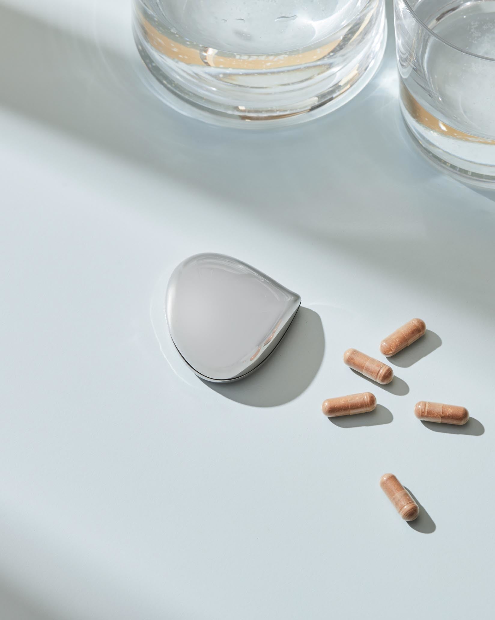 Polished silver chestnut-shaped object by Alessi, displayed with brown capsules and two water glasses on a blue surface.