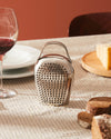 Silver bell-shaped grater by Alessi with a handle, surrounded by grated cheese on a checkered tablecloth.