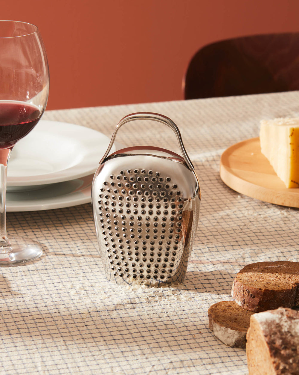 Silver bell-shaped grater by Alessi with a handle, surrounded by grated cheese on a checkered tablecloth.