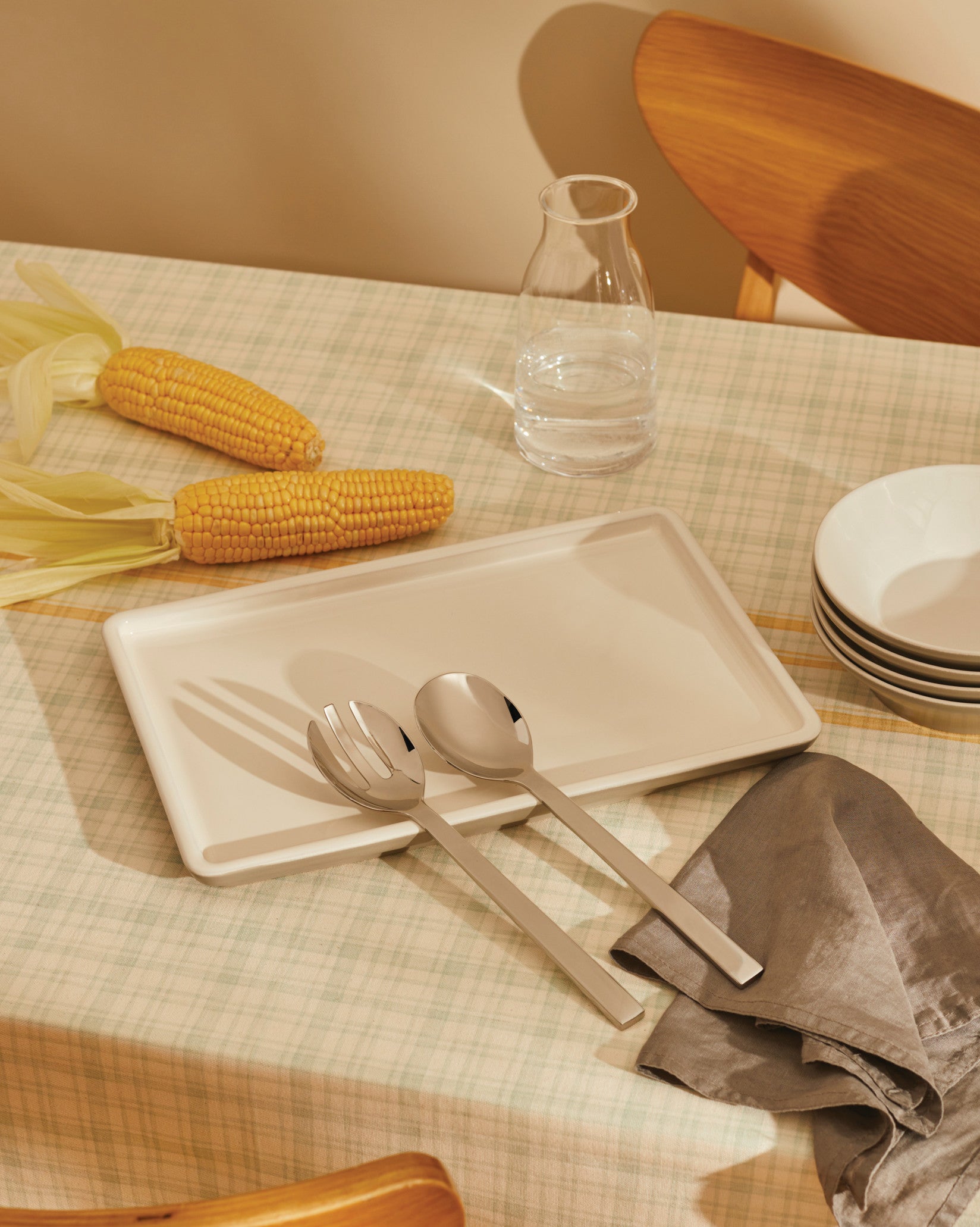Off-white ceramic serving platter by Alessi, holding silver utensils on a plaid tablecloth with corn and bowls.