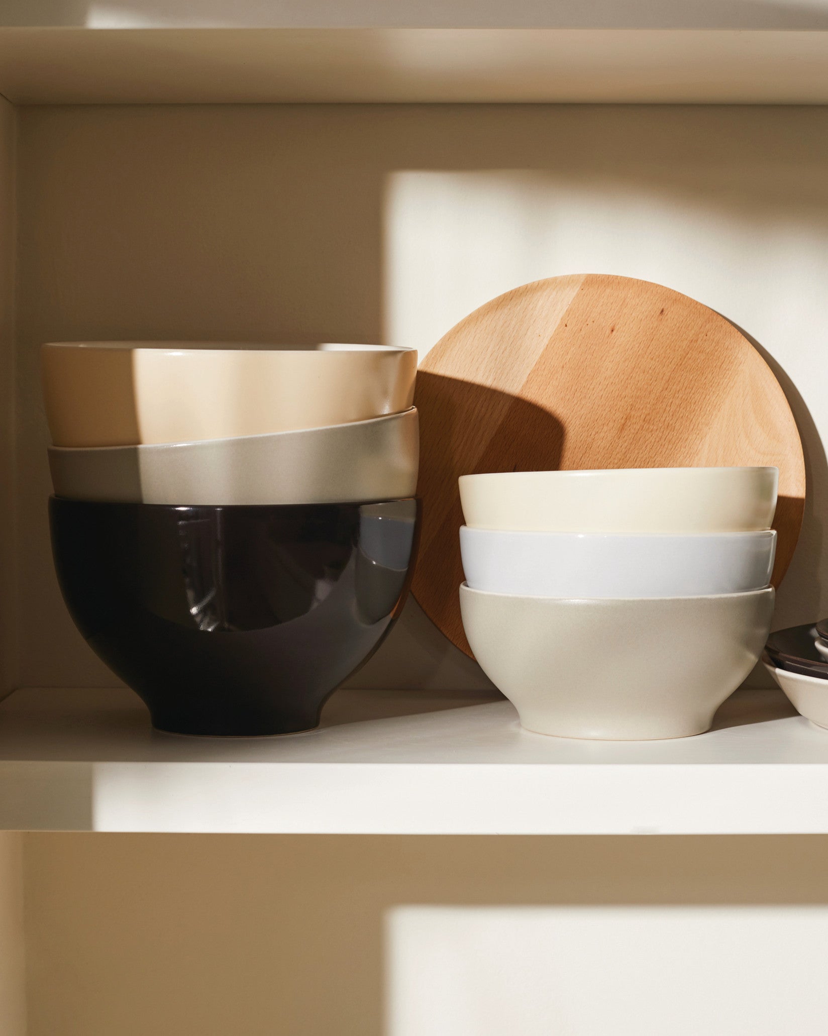 Tonale ceramic bowls in varying neutral colors by Alessi, stacked on a white shelf with a round wooden board.