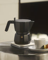 Black matte faceted moka pot by Alessi with a gold safety valve, on a coaster beside a coffee cup.