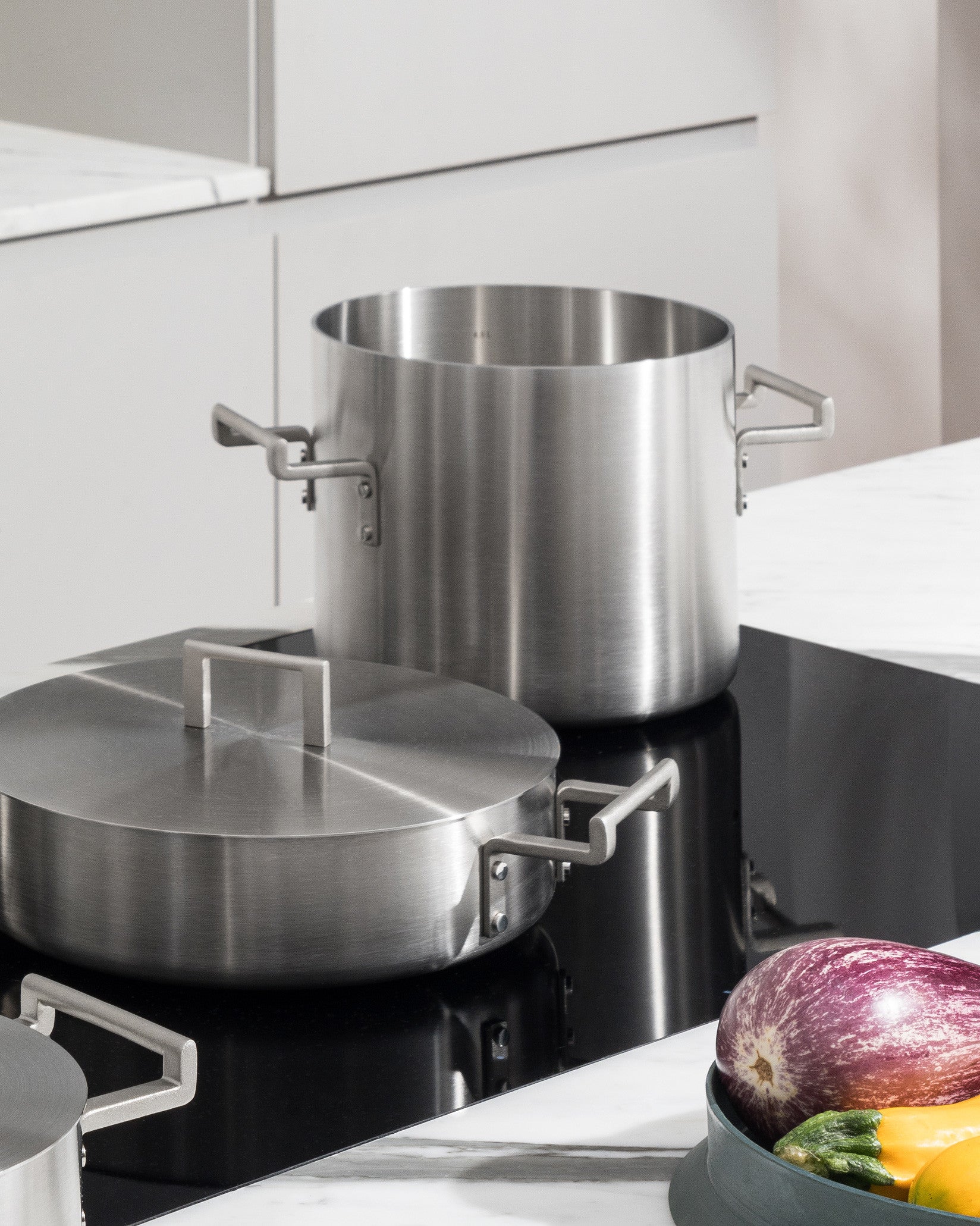 Two brushed stainless steel pots by Alessi, one lidded, with minimalist handles, on a black induction stovetop.