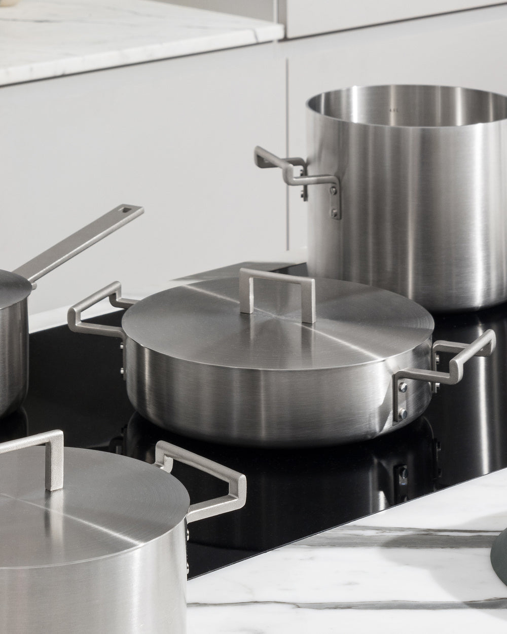 Stainless steel pots from the Convivio collection by Alessi, with minimalist handles, on a black stovetop in a modern kitchen.