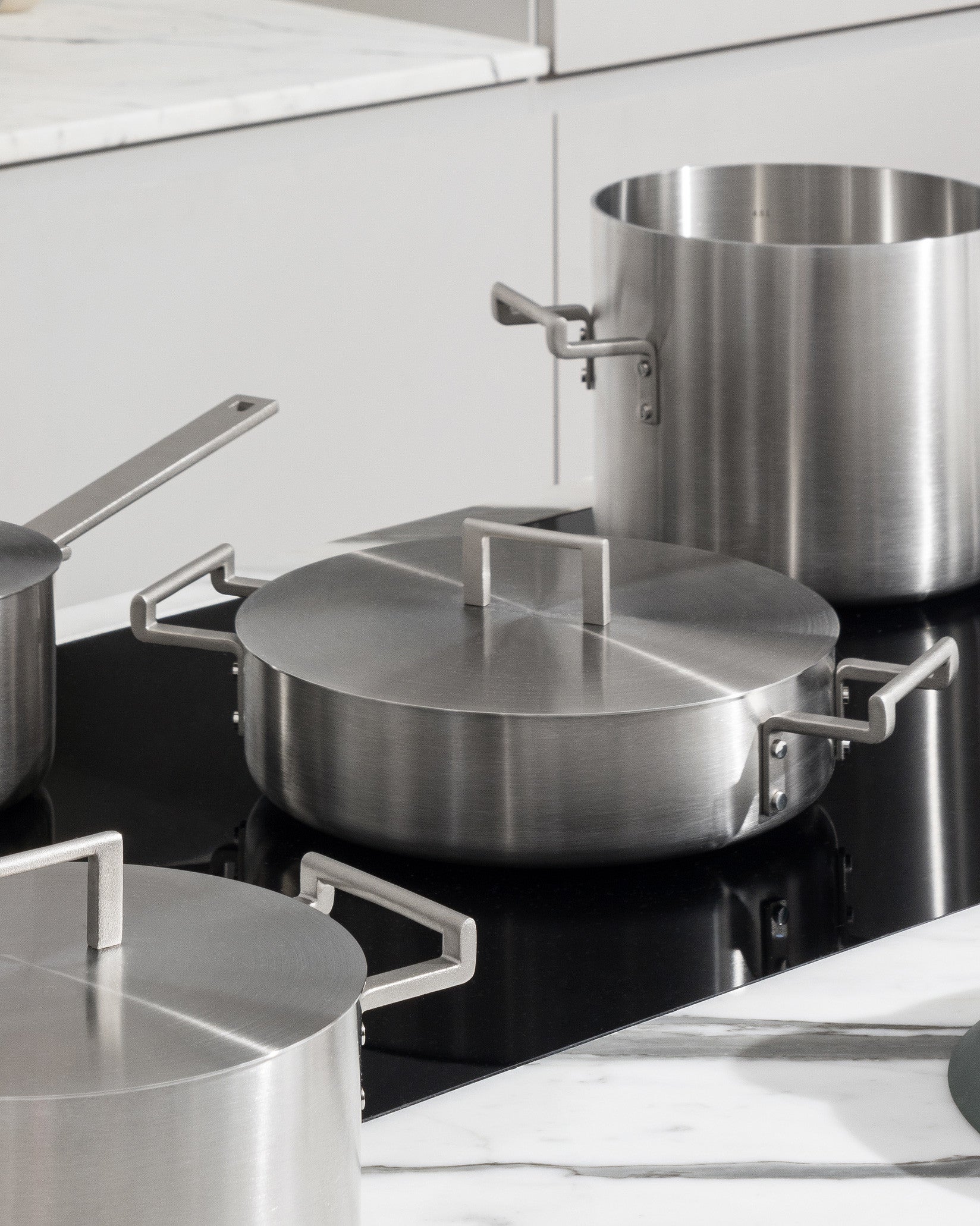 Stainless steel pots from the Convivio collection by Alessi, with minimalist handles, on a black stovetop in a modern kitchen.