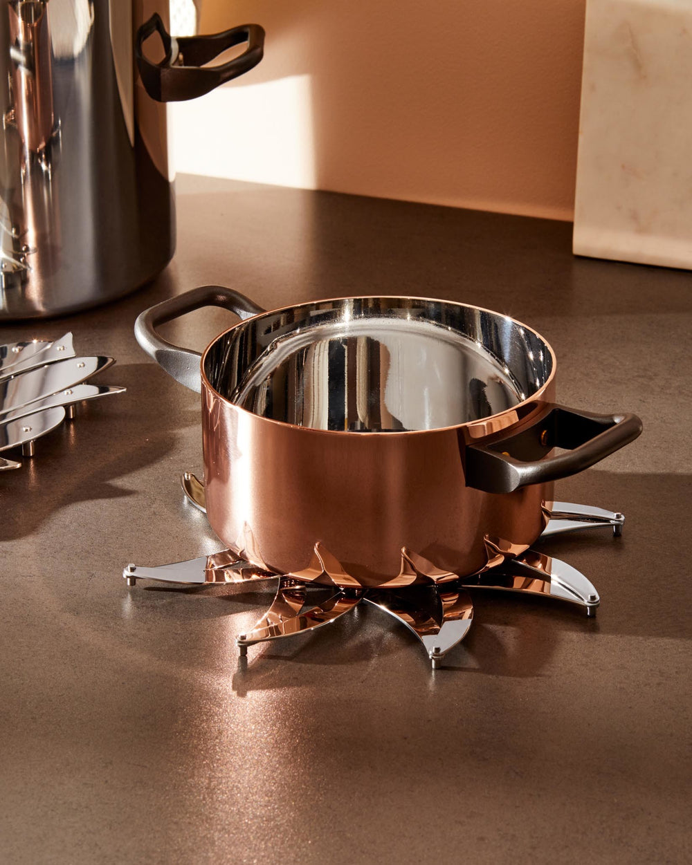 Shiny silver-toned metal trivet by Alessi featuring an expandable petal design, supporting a copper pot on a dark countertop.