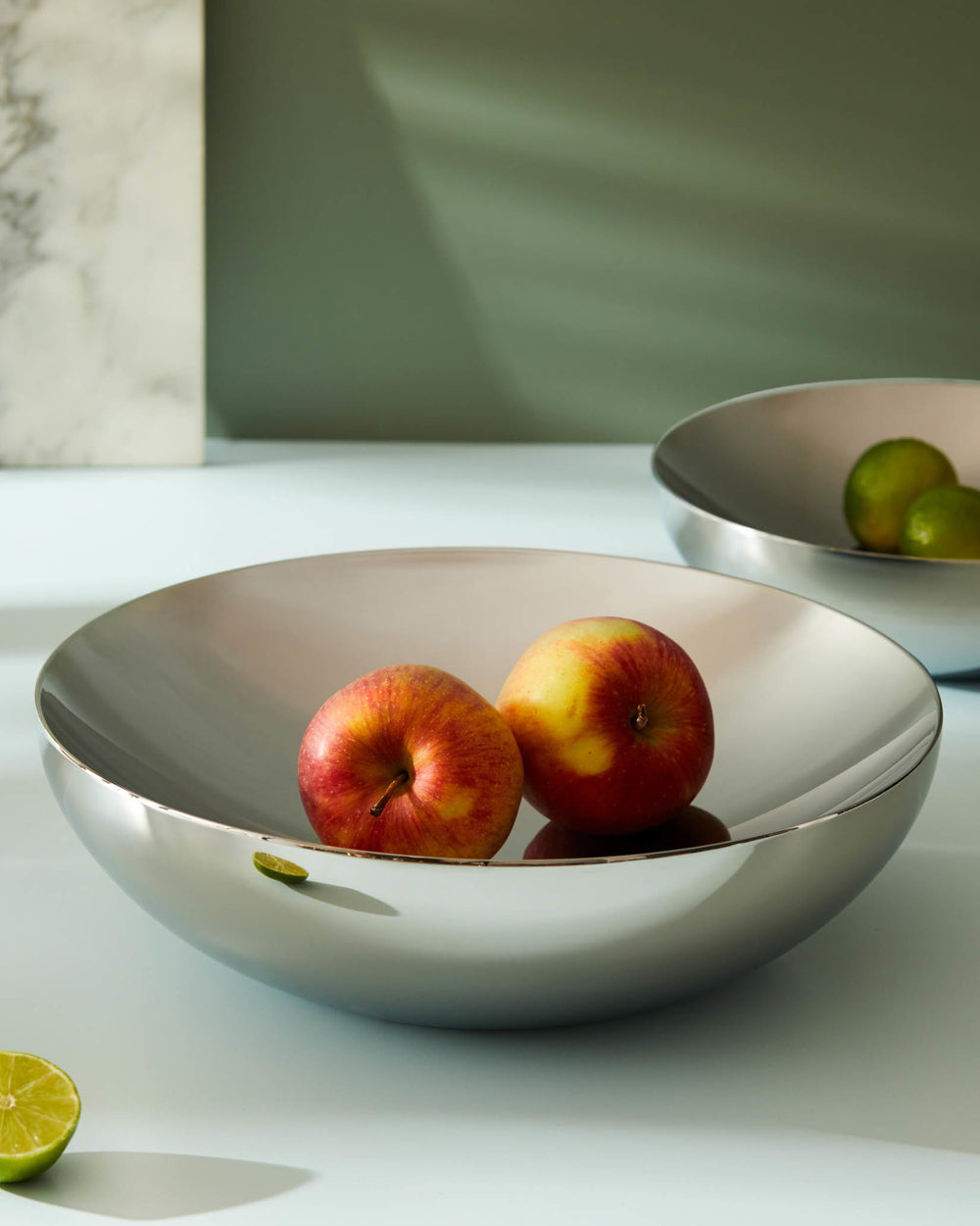 Sleek silver Double fruit bowl by Alessi, holding two apples and a lime slice, with a blurred background bowl.