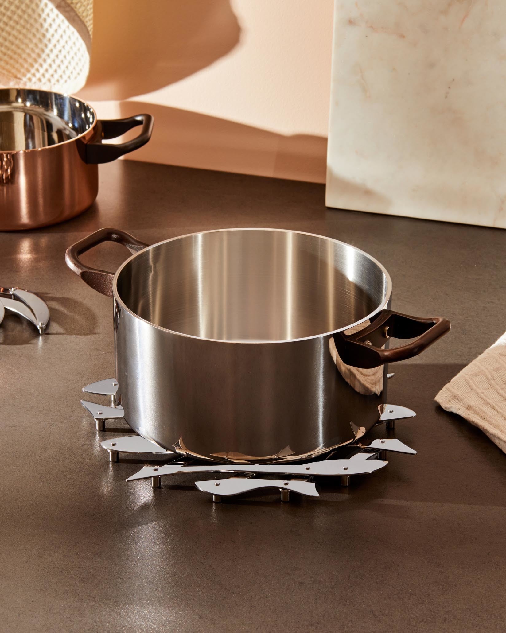 Silver-toned metal trivet by Alessi, designed with interlocking fish shapes supporting a large cooking pot on a dark counter.