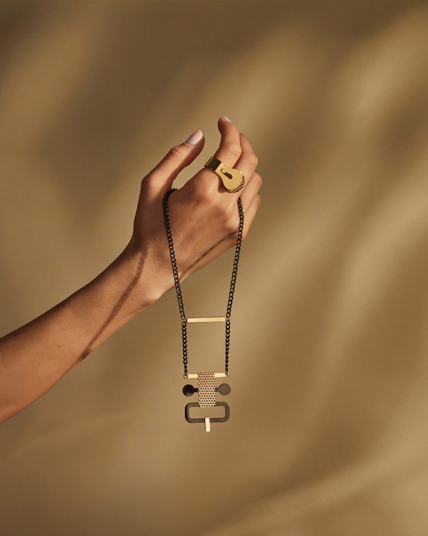 Edone geometric pendant necklace by Alessi with a dark chain, featuring gold and patterned details, held by a hand wearing a wide gold ring.
