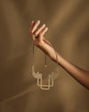 Textured gold-toned geometric necklace by Alessi, held by a hand wearing a patterned ring against a brown background.
