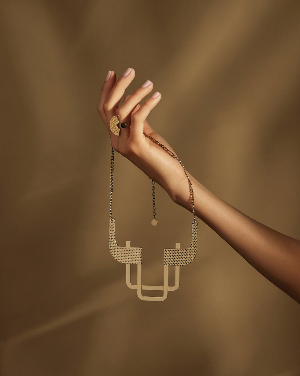 Textured gold-toned geometric necklace by Alessi, held by a hand wearing a patterned ring against a brown background.