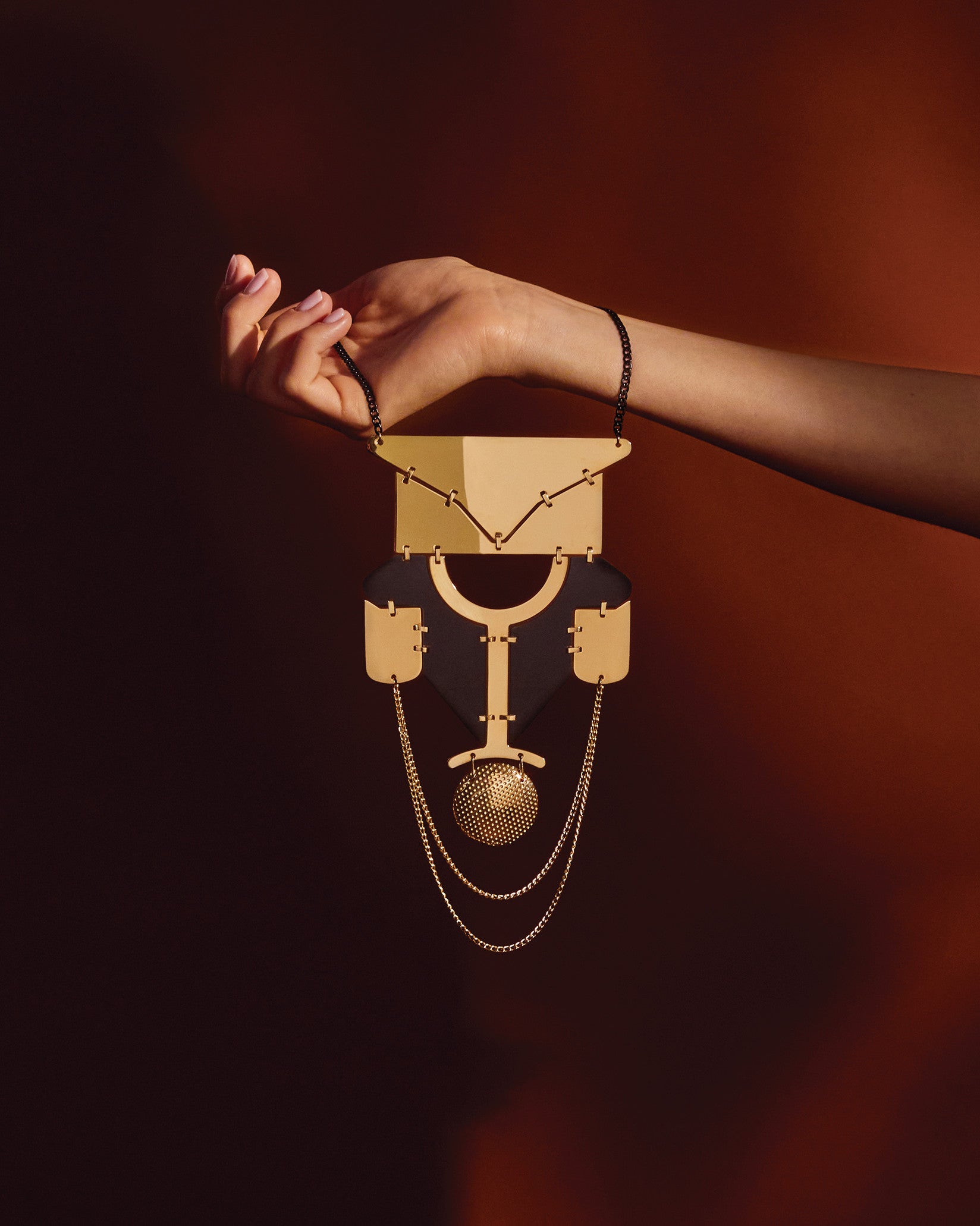 Gold and black geometric pendant necklace by Alessi, with a textured sphere, held by a hand.