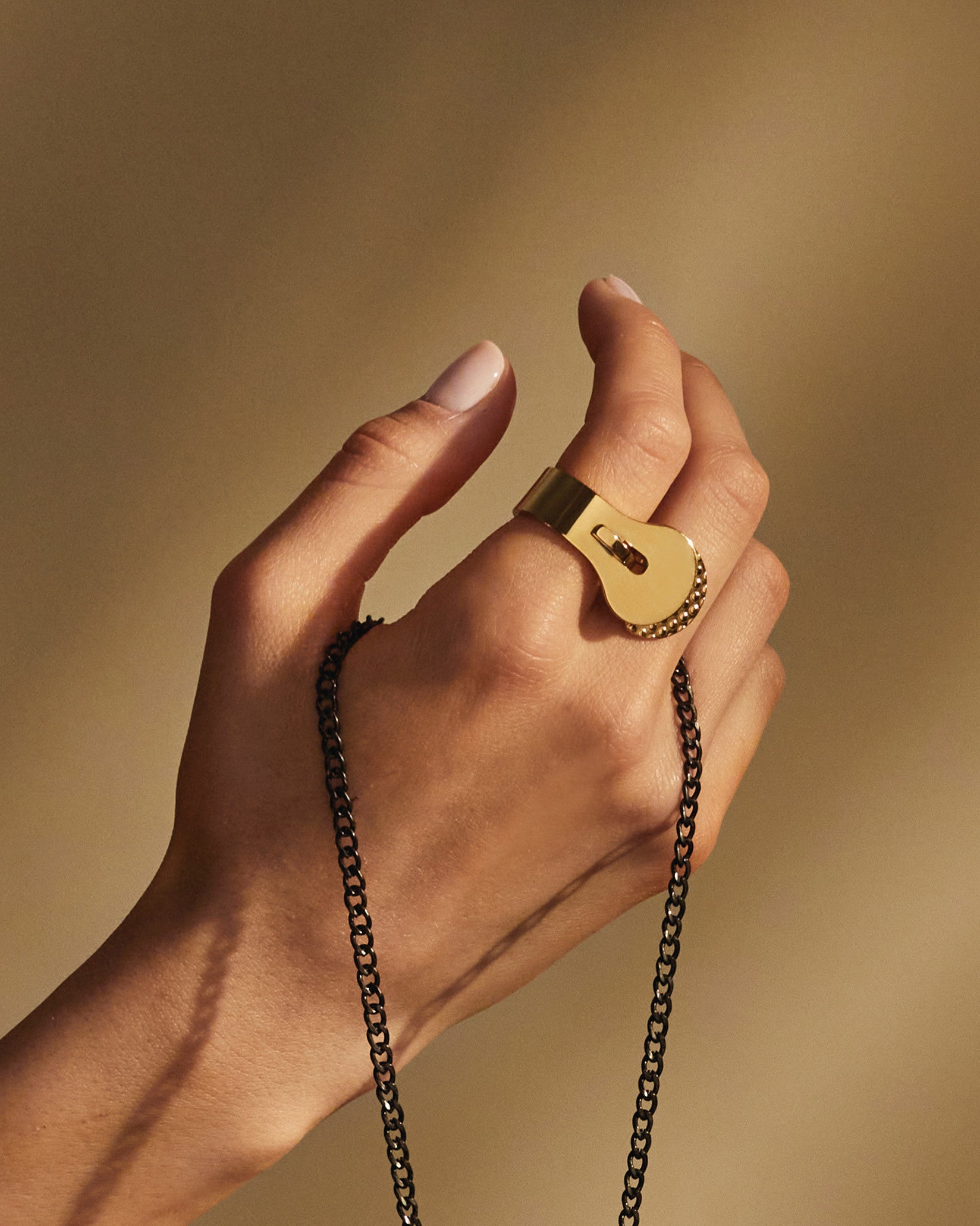 Golden Trama ring by Alessi, featuring a lock-like design with a studded edge, worn on a hand holding a dark chain.