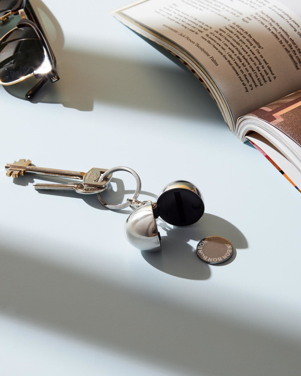 Open silver spherical keychain container by Alessi, revealing a black interior, with a 'BON BON' coin nearby.