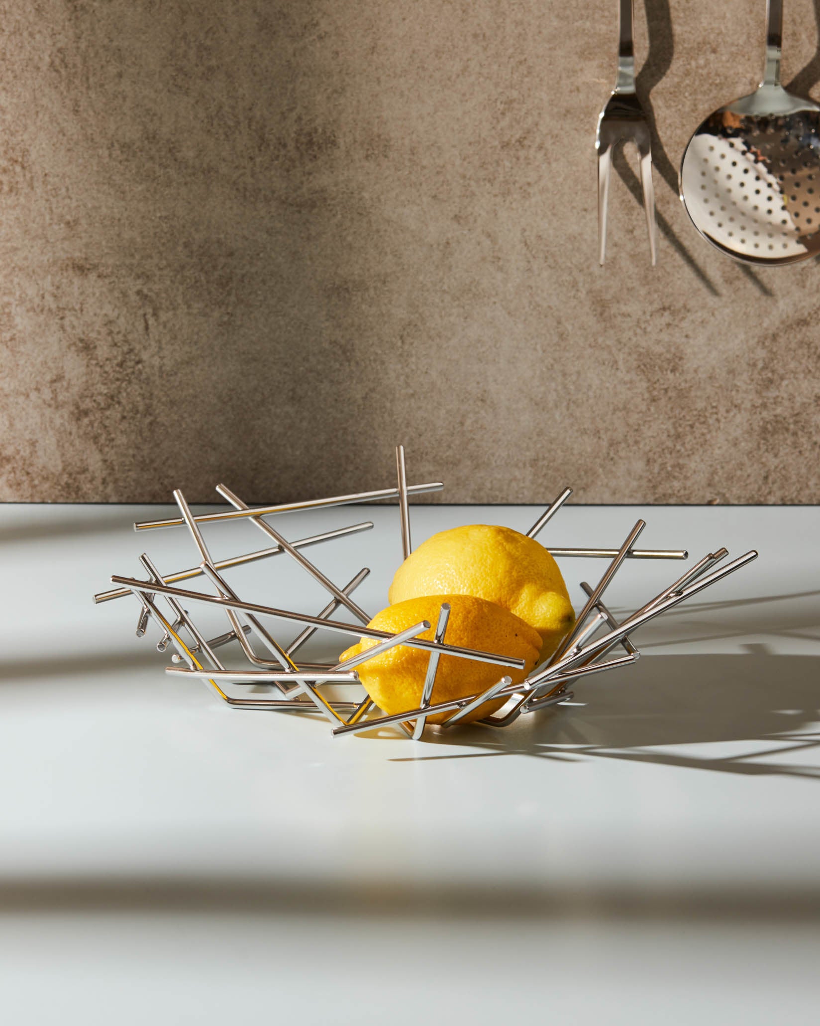 Modern stainless steel fruit basket by Alessi, with a geometric stick design, holding two lemons on a white counter.