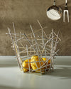 Silver metal fruit bowl by Alessi with an abstract, crisscrossing rod design, holding yellow lemons on a counter with kitchen utensils.