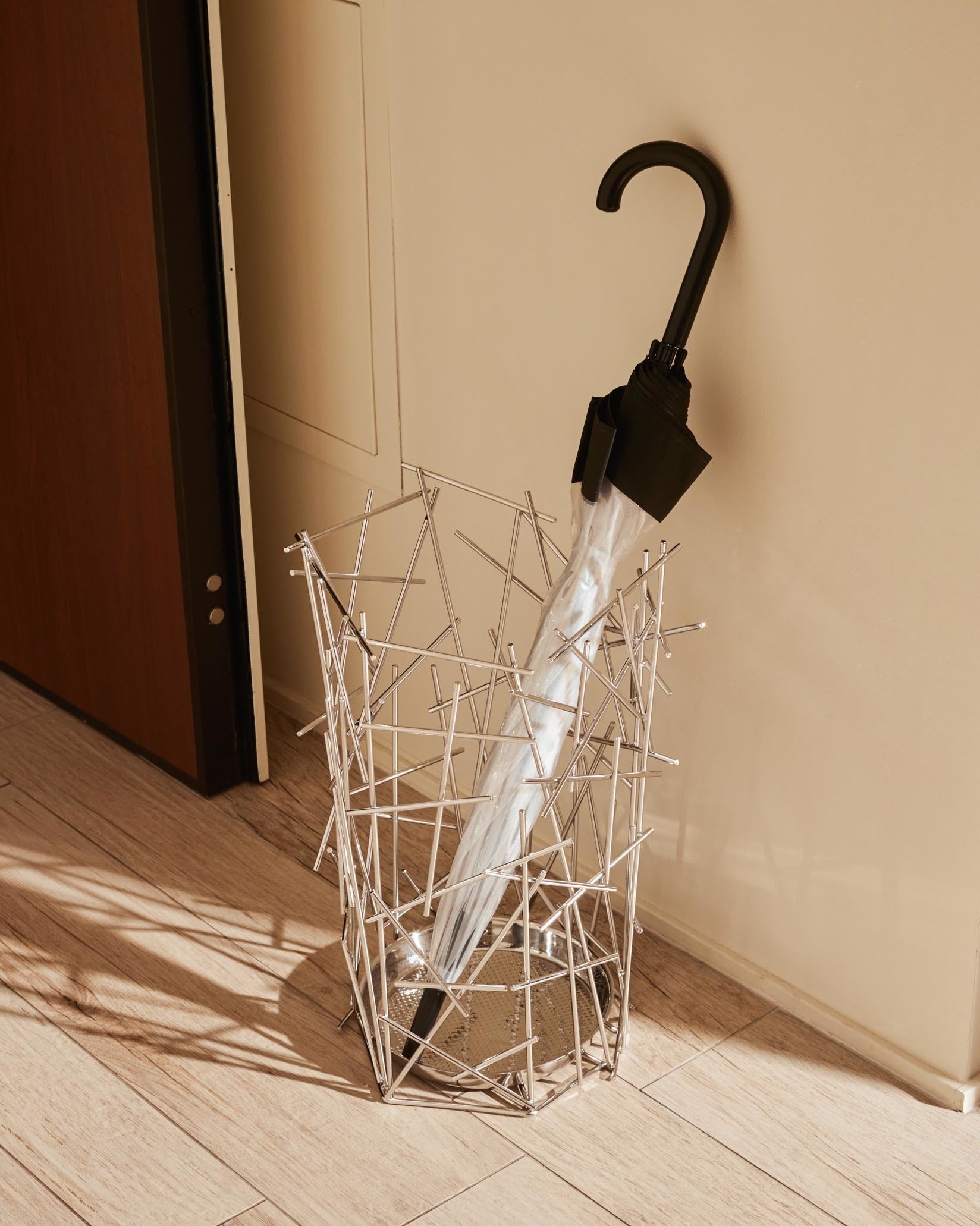 Silver metal umbrella stand by Alessi, featuring a geometric crisscross rod design with a black umbrella inside on a wooden floor.