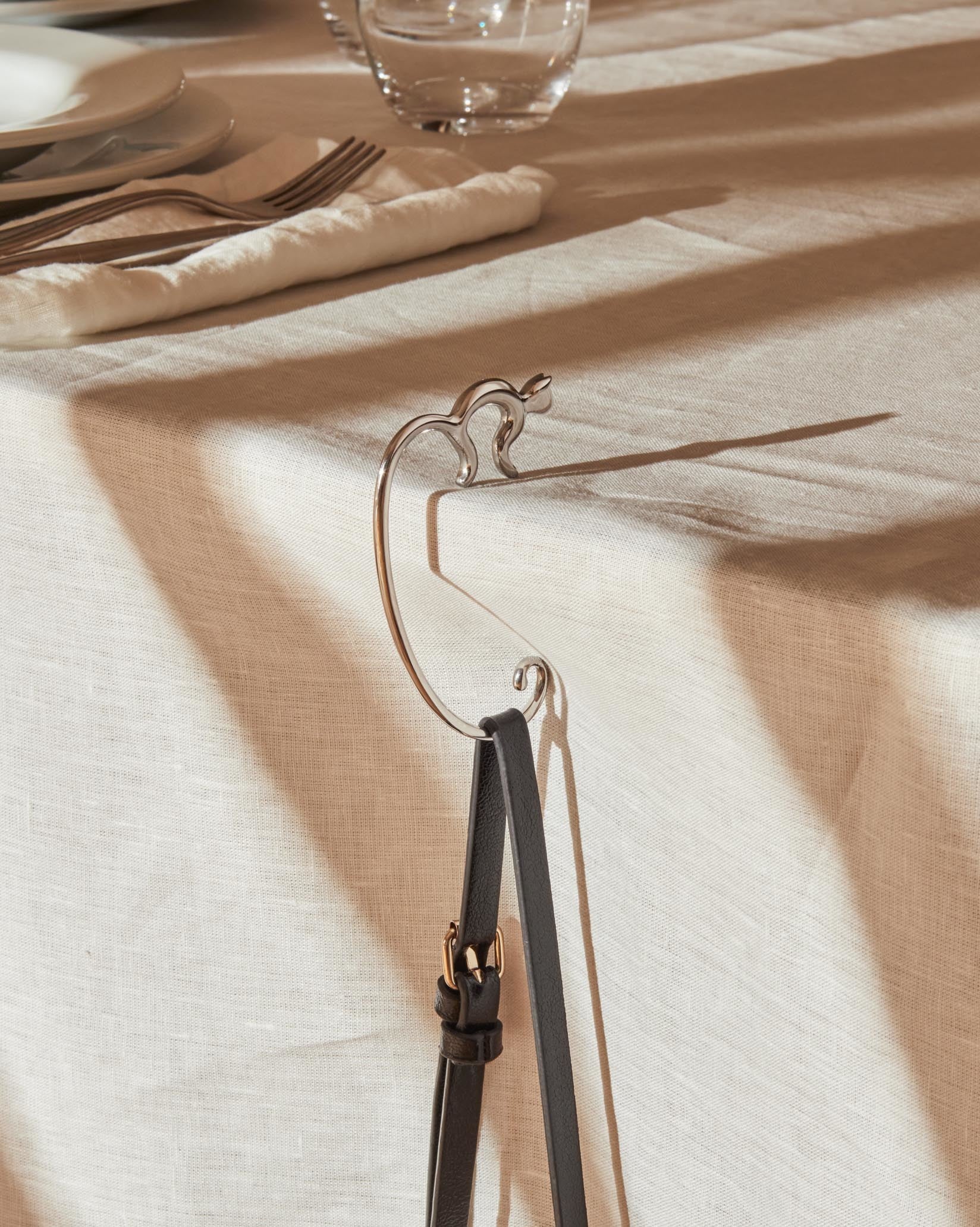 Silver-toned metal handbag hook by Alessi, cat-shaped, holding a black leather bag strap on a linen tablecloth.