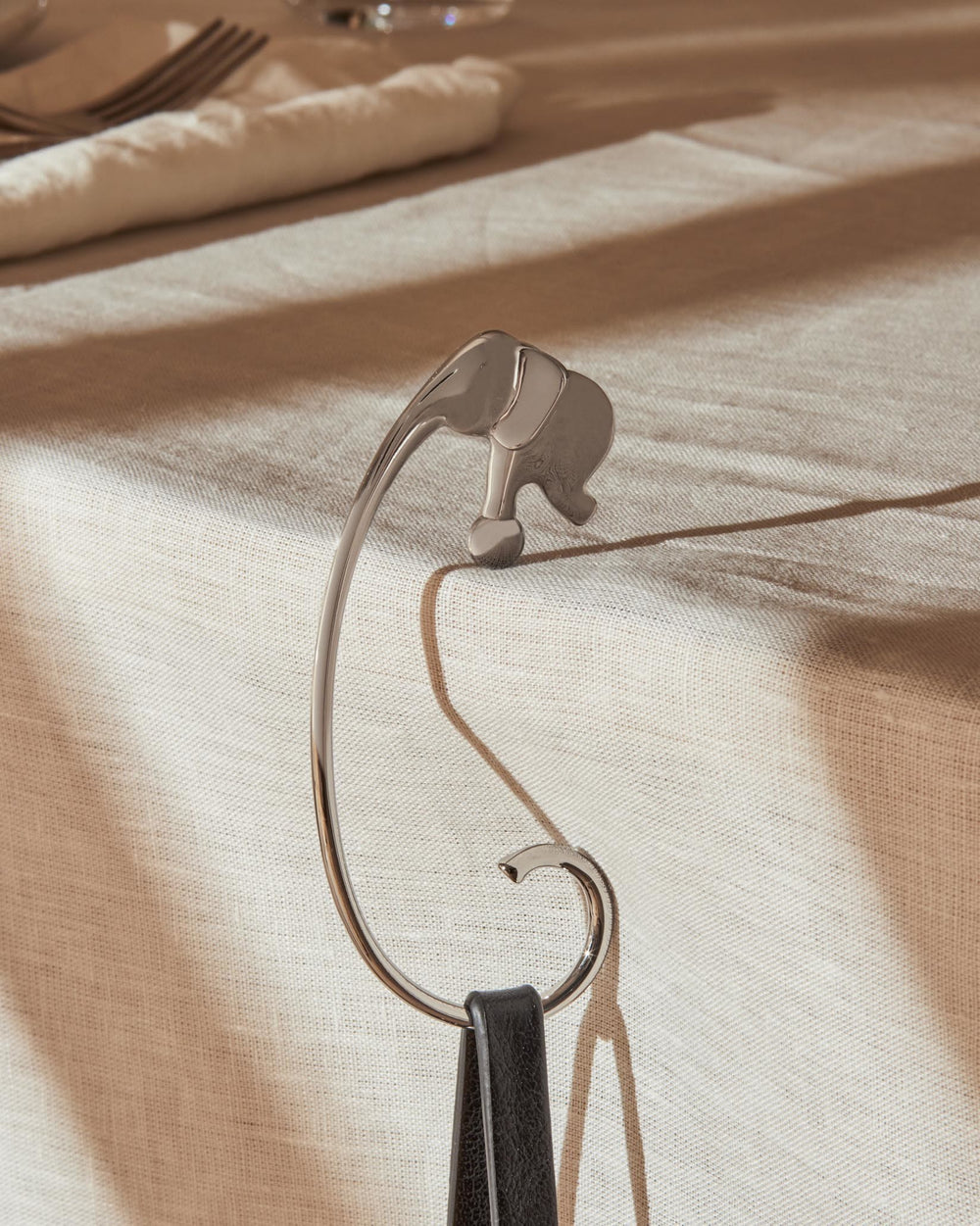 Silver-toned elephant bag hook by Alessi, hanging from a table edge with a black strap on a linen tablecloth.