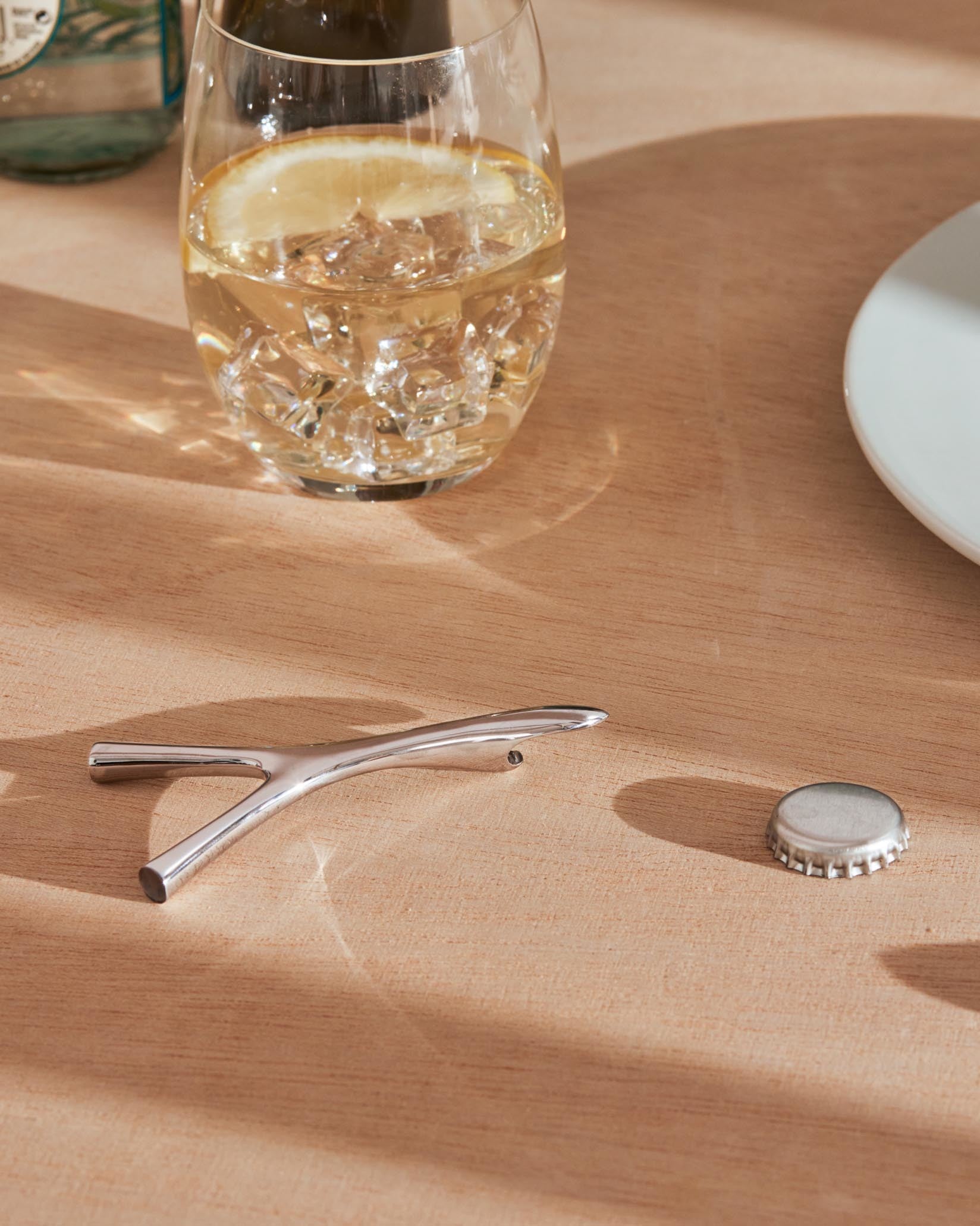 Silver metal bottle opener by Alessi, with a Y-shaped design, on a wooden table next to a bottle cap.