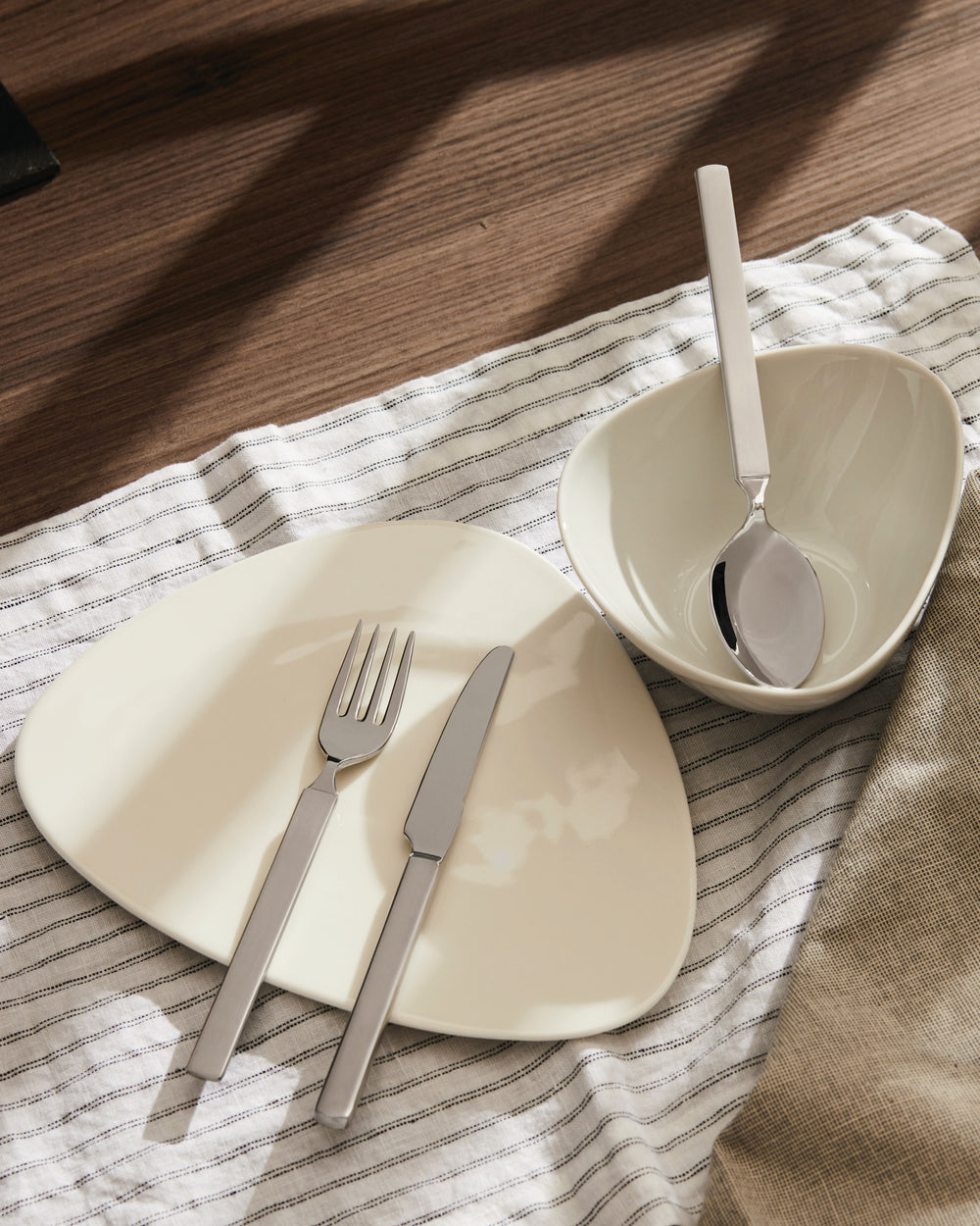 Modern flat-handled silver cutlery and off-white dishes by Alessi, displayed with striped napkins on a wood table.