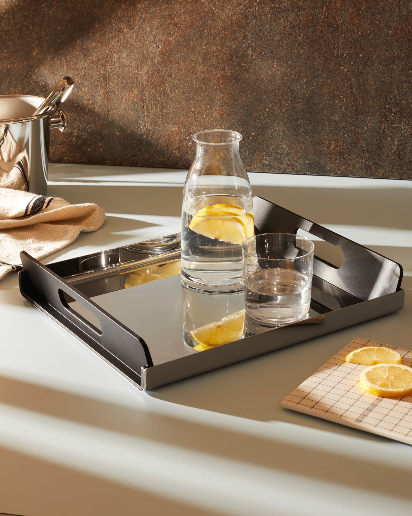 A dark grey and polished stainless steel serving tray by Alessi, on a light blue countertop, holding a carafe and glass of lemon water.